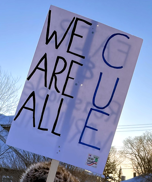 photo: picketer with sign which reads, 'WE ARE ALL CUE'